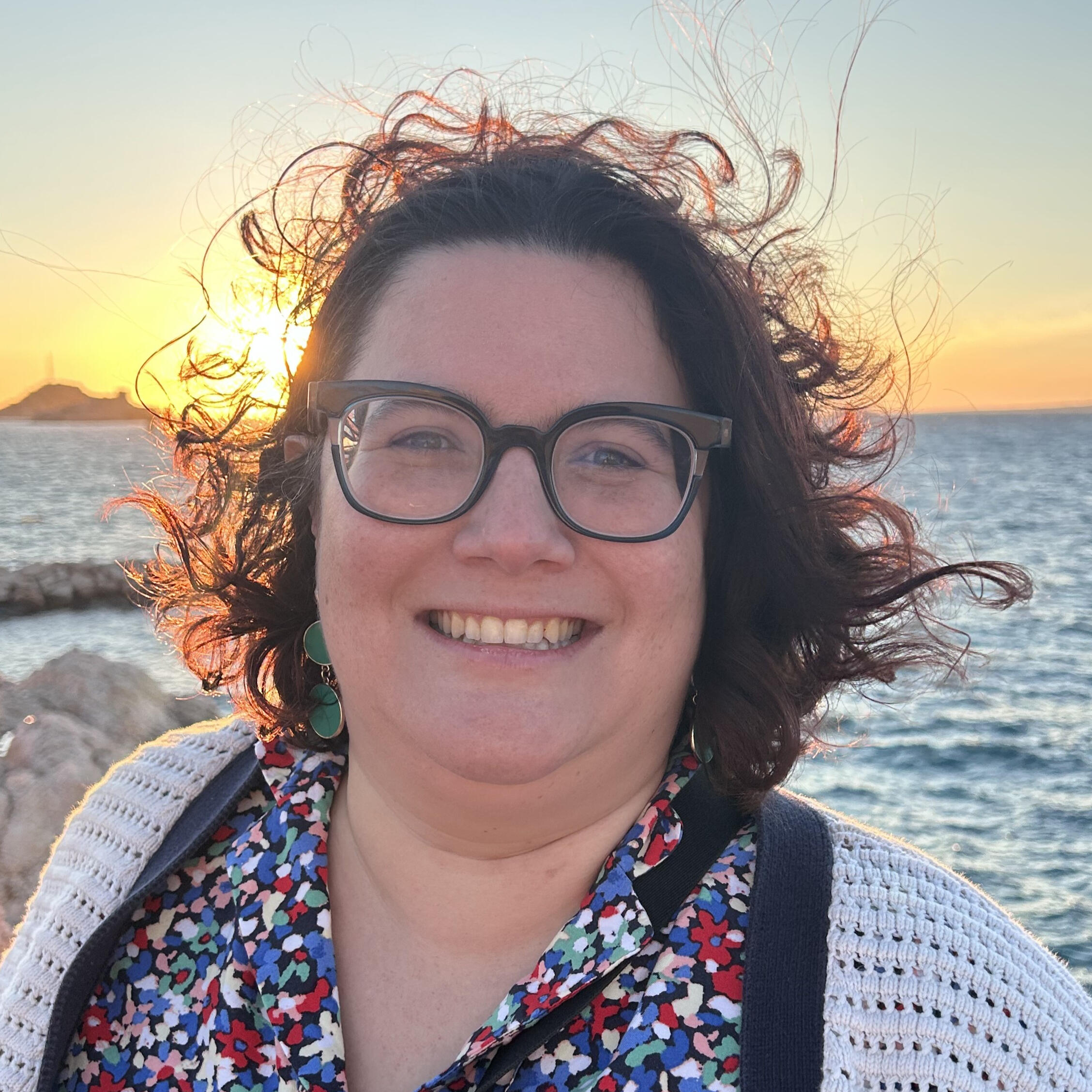 a female (Sarah Nolasco) with glasses with a sunset behind her. She has on a floral dress with red, white and blue flowers.