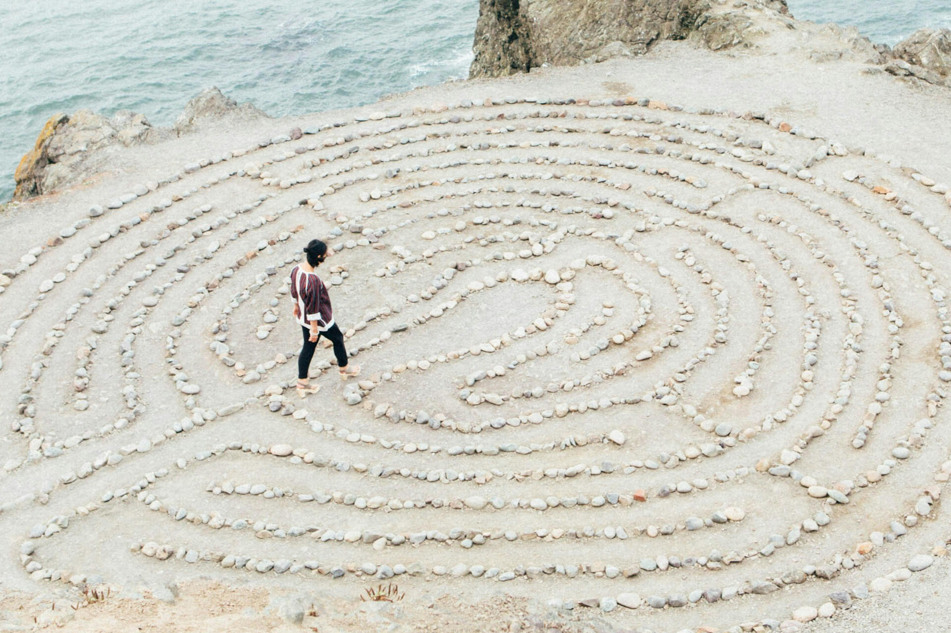 a women walking in a labyrinth by the sea. The walls are low and made of rocks